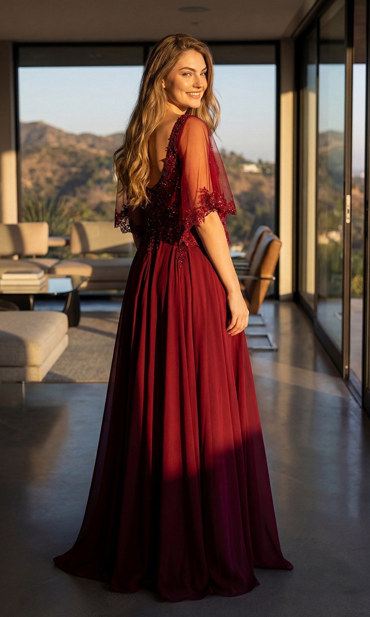 A woman stands indoors wearing the Juliet JTM22U long formal dress in burgundy, featuring delicate lace details and an embroidered sheer bodice. She looks over her shoulder, with modern furniture and large windows visible in the background.